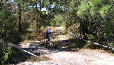 A cyclist in a blue helmet rides along a rocky trail surrounded by trees and greenery. The path is marked with tape, indicating a specific route for biking. Georgia International Horse Park mountain bike trail.