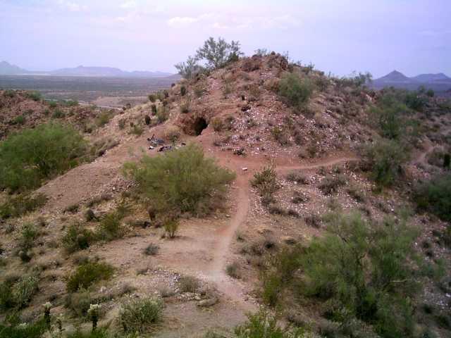 A rocky hill surrounded by desert vegetation, including scattered shrubs and small trees. A winding dirt path leads up the hill, with distant mountain ranges visible in the background under a cloudy sky. The landscape features a variety of earthy tones and textures typical of a dry environment. Hawes Loop mountain bike trail.