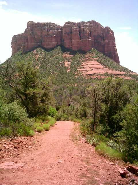 A dirt path leads through lush green vegetation toward a prominent rock formation with steep, layered sides under a clear blue sky. Hawes Loop mountain bike trail.