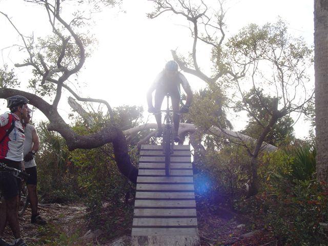 A mountain biker is mid-jump off a wooden ramp, surrounded by dense foliage and trees. Another cyclist in the foreground watches the jump, which takes place in a natural outdoor setting. The sunlight creates a bright background effect, highlighting the action of the jump. Jonathan Dickinson State Park mountain bike trail.