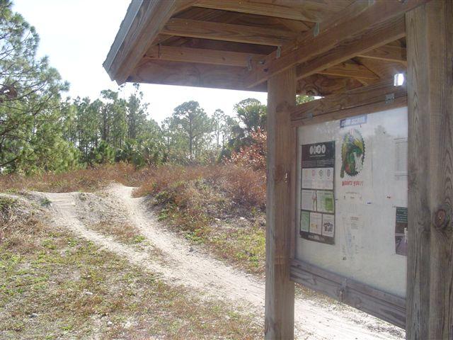 A wooden informational sign at a trailhead, displaying maps and guidelines, beside a winding dirt path leading into a wooded area with sparse vegetation and trees in the background. Jonathan Dickinson State Park mountain bike trail.