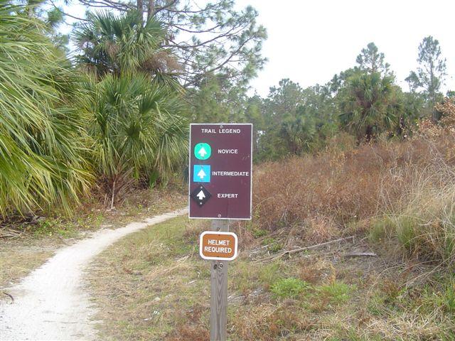 A trail sign indicating difficulty levels: novice, intermediate, and expert, along with a notice that a helmet is required. The sign is situated along a winding dirt path surrounded by trees and vegetation. Jonathan Dickinson State Park mountain bike trail.