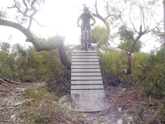 Mountain biker performing a jump off a wooden ramp in a wooded area, surrounded by trees and foliage. Jonathan Dickinson State Park mountain bike trail.