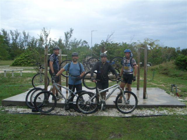 Four male mountain bikers pose together on a grassy area, with their bicycles nearby. They are wearing helmets and cycling attire. In the background, there are trees and a cloudy sky, suggesting an outdoor cycling location. The group appears to have stopped for a moment during their ride. Oleta River State Park mountain bike trail.