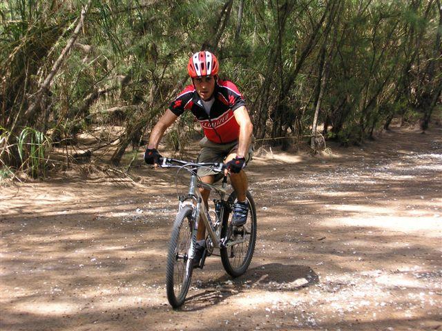 A person riding a mountain bike on a dirt trail surrounded by trees, wearing a red helmet and a red and black cycling jersey. The scene is well-lit with dappled sunlight filtering through the foliage. Oleta River State Park mountain bike trail.