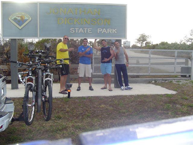 Four men are gathered in front of the Jonathan Dickinson State Park sign, smiling and posing with thumbs up. Two bicycles are parked in the foreground, and the landscape features sparse greenery typical of a Florida park. The scene suggests a day of outdoor activity and camaraderie. Jonathan Dickinson State Park mountain bike trail.