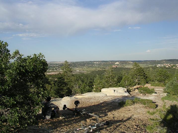 A person sitting on a rocky outcrop overlooking a vast landscape of rolling hills and evergreen trees, with a clear blue sky above. A bicycle is resting on the ground nearby. Ute Valley Park mountain bike trail.