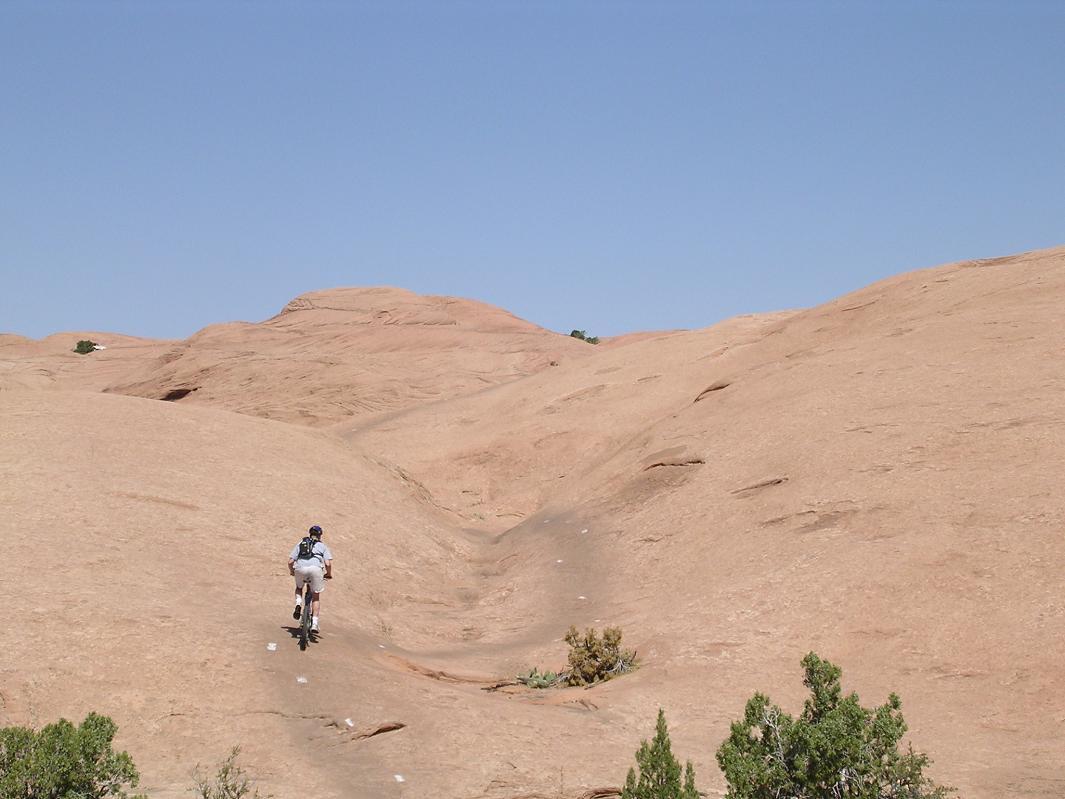 A mountain biker ascends a rocky, desert landscape with smooth, reddish-brown terrain under a clear blue sky. Sparse vegetation, including small bushes, is visible along the trail. The path is marked with white dots leading into the distance. Slickrock mountain bike trail.