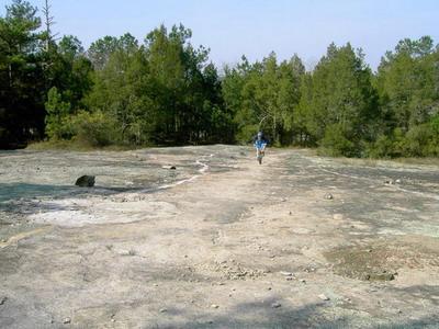 A person walking on a rocky, uneven terrain surrounded by trees in a natural setting. The landscape features patches of bare ground and sparse vegetation, with the figure moving away from the camera toward the tree line. Georgia International Horse Park mountain bike trail.