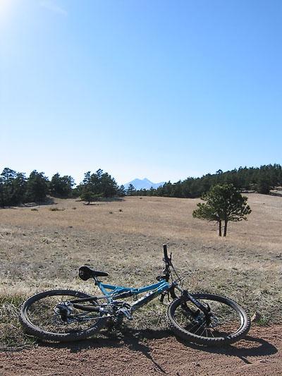A mountain bike lying on a dirt path in an open field, with distant mountains and clear blue skies in the background. Surrounding trees and grass create a natural landscape. Hall Ranch mountain bike trail.