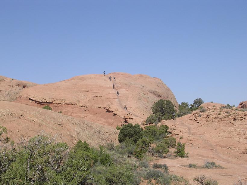 A rocky landscape featuring a group of hikers ascending a large, smooth, reddish-brown rock formation under a clear blue sky. Green shrubs and small trees are scattered throughout the foreground, enhancing the natural scenery. Slickrock mountain bike trail.