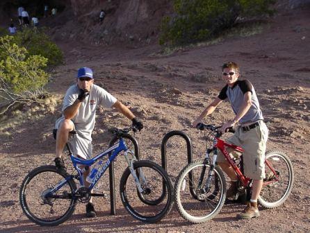 Two young men, one sitting on a blue mountain bike and the other standing next to a red mountain bike, pose at a bike rack in a rocky outdoor setting. Both are dressed in casual clothing suitable for biking, with green shrubbery and a cliff in the background. The scene captures a moment of relaxation and camaraderie amid a biking adventure. Papago Park Area mountain bike trail.