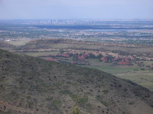 A panoramic view of rolling hills and rugged terrain, with a distant city skyline visible in the background. The landscape features a mix of green vegetation and reddish rock formations, against a clear blue sky. Deer Creek Canyon mountain bike trail.