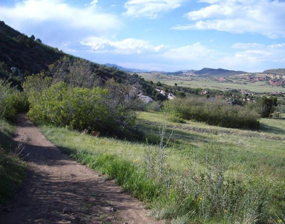 A scenic landscape featuring a dirt path winding through a grassy field, bordered by shrubs and trees. In the background, rolling hills and mountains stretch under a bright blue sky with scattered clouds. Deer Creek Canyon mountain bike trail.