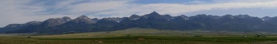 Panoramic view of a mountain range with snow-capped peaks, set against a clear blue sky, above a broad green valley. Rainbow Trail: Westcliffe mountain bike trail.