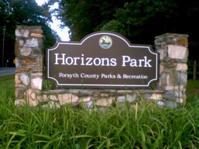 Sign for Horizons Park, featuring a brown background with white text that reads "Horizons Park" and "Forsyth County Parks & Recreation," surrounded by natural stone and greenery. Horizons Park mountain bike trail.