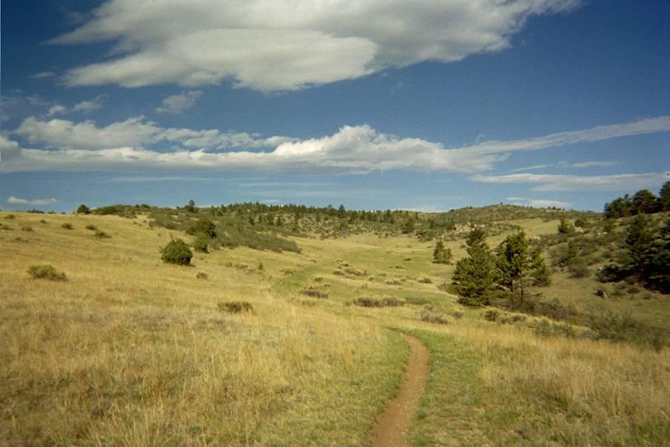 A winding dirt path leading through a grassy landscape, surrounded by rolling hills and patches of trees under a blue sky with scattered clouds. Hewlett Gulch mountain bike trail.