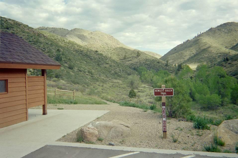 A scenic view of a hiking trailhead in a mountainous area. In the foreground, there is a wooden structure with a brown exterior. A trail sign indicating the "Hewlett Gulch Trail" is visible next to a well-marked path leading into the lush green valley and rolling hills. The sky is partly cloudy, and the surrounding landscape is dotted with trees and rocky outcrops. Hewlett Gulch mountain bike trail.