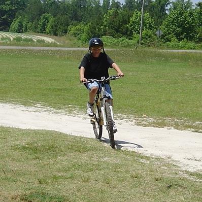 A young person wearing a helmet rides a bicycle on a sandy path in a grassy outdoor area, surrounded by trees. San-lee Park mountain bike trail.