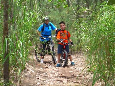 A woman and a young boy stand beside their mountain bikes on a dirt path surrounded by tall grass and trees, enjoying a moment in nature. The woman is wearing a blue shirt and a helmet, while the boy is dressed in an orange shirt and shorts. Oleta River State Park mountain bike trail.