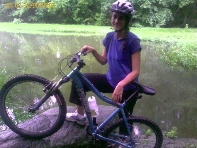 A woman wearing a helmet and casual biking attire sits on a rock beside a river, with a mountain bike next to her. She has a water bottle attached to the bike and smiles as she poses for the photo, surrounded by lush green foliage. Salem Lake mountain bike trail.