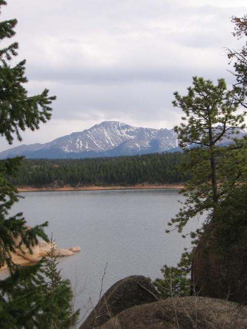 A scenic view of a mountain with snowy peaks in the background, framed by pine trees, overlooking a calm lake. The sky is cloudy, and a lush forest surrounds the water's edge. Rampart Reservoir mountain bike trail.
