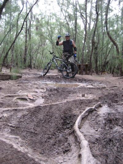 A person standing next to a mountain bike on a muddy trail surrounded by trees. The rider is wearing a helmet, gloves, and a sleeveless shirt, and is giving a thumbs up. Visible roots and dirt marks indicate mud and trail activity. Oleta River State Park mountain bike trail.