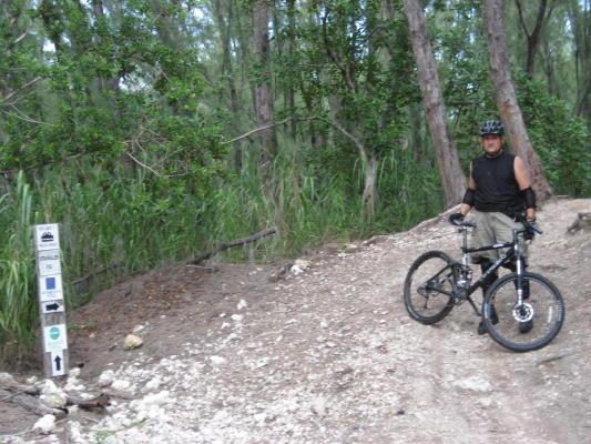 A person standing next to a mountain bike on a dirt trail surrounded by trees and greenery. There is a trail sign indicating directions and difficulty levels in the foreground. The individual is wearing a helmet and a sleeveless shirt, ready to embark on a biking adventure. Oleta River State Park mountain bike trail.