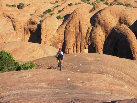 A mountain biker riding down a rocky terrain surrounded by unique rock formations and vegetation under a clear blue sky. Slickrock mountain bike trail.