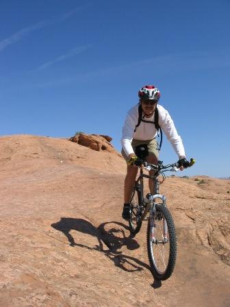 A person riding a mountain bike on a rocky terrain under a clear blue sky. The cyclist is wearing a helmet and athletic clothing, navigating the uneven surface with determination. Slickrock mountain bike trail.