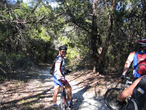 A mountain biker in a blue and white outfit stands on a forest trail, looking back at the camera. Surrounding them are tall trees, with dappled sunlight filtering through the foliage. Another cyclist can be seen in the foreground, partially obscured, as they navigate the path. Cameron Park mountain bike trail.