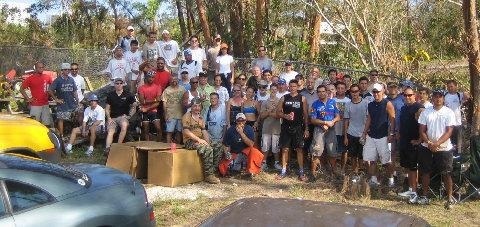 A large group of people gathered outdoors, with a mix of men and women of diverse backgrounds. They are posing together in a casual setting, with some seated on boxes and others standing behind them. The background features trees and a fence, indicating a community or social event. Oleta River State Park mountain bike trail.