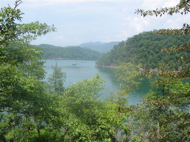 A serene view of a calm lake surrounded by lush green trees and rolling hills under a partly cloudy sky. The reflection of the mountains is visible on the water's surface, creating a tranquil landscape. Tsali Left Loop mountain bike trail.