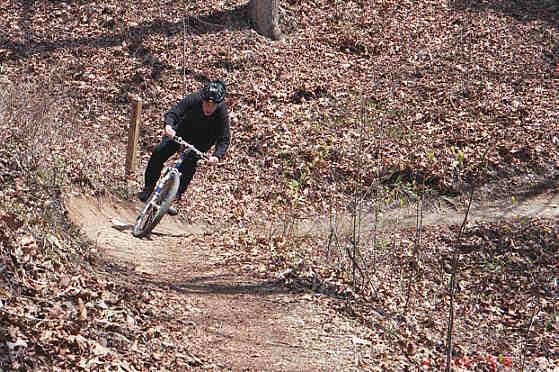 A person in a black outfit rides a bicycle along a winding dirt trail surrounded by fallen leaves and sparse vegetation in a wooded area. Tsali Left Loop mountain bike trail.