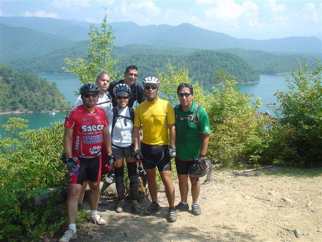 A group of six cyclists posing for a photo on a trail with a scenic view of a lake and mountains in the background. They are wearing various cycling jerseys and helmets, with green foliage surrounding them. The sky is partly cloudy, indicating a beautiful day for biking. Tsali Mouse Branch Loop mountain bike trail.