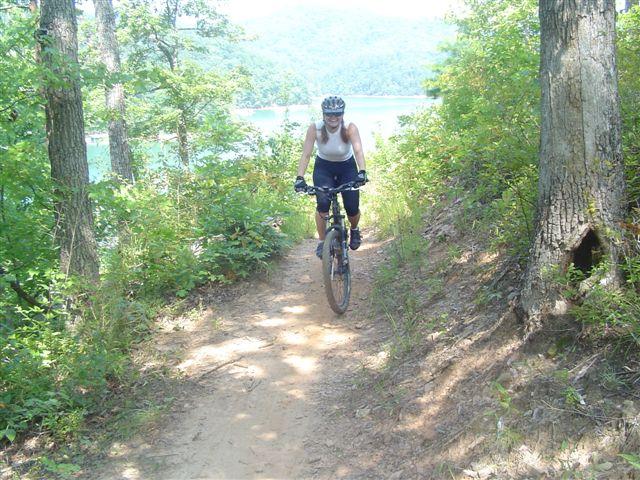 A person riding a mountain bike on a dirt trail surrounded by greenery, with a lake and mountains visible in the background. The cyclist is wearing a helmet and athletic clothing, enjoying a sunny day outdoors. Tsali Mouse Branch Loop mountain bike trail.