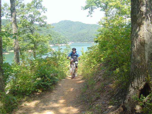 A mountain biker riding along a narrow dirt path surrounded by lush greenery, with a serene lake and rolling hills visible in the background under a clear blue sky. Tsali Mouse Branch Loop mountain bike trail.