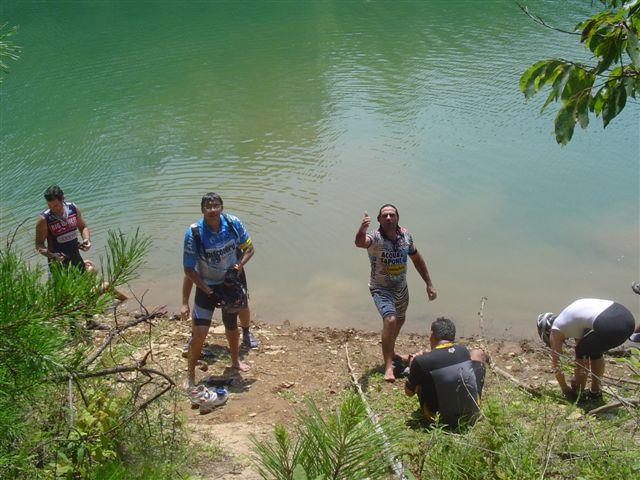 A group of four individuals is gathered by a body of water, smiling and interacting with each other. Two men are standing on the shore, one appears to be holding something and gesturing playfully, while a third man crouches down near the water's edge. The fourth person is partially visible, focused on something on the ground. The setting is lush and green, with trees surrounding the area, indicating a natural outdoor environment. Tsali Mouse Branch Loop mountain bike trail.