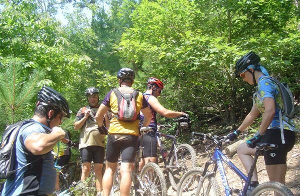 A group of mountain bikers gathered on a trail surrounded by lush greenery. Some riders are adjusting their gear while others are engaged in conversation, with several mountain bikes resting nearby. The sun is shining through the trees, creating a vibrant outdoor atmosphere. Tsali Mouse Branch Loop mountain bike trail.