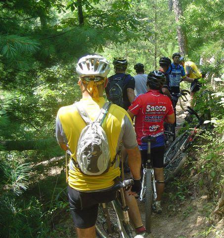 A group of mountain bikers is navigating a narrow trail through a lush, green forest. Some riders are wearing brightly colored jerseys, and they are positioned along the path, with their bikes parked beside them. The scene captures the excitement and camaraderie of outdoor cycling in a natural setting. Tsali Mouse Branch Loop mountain bike trail.