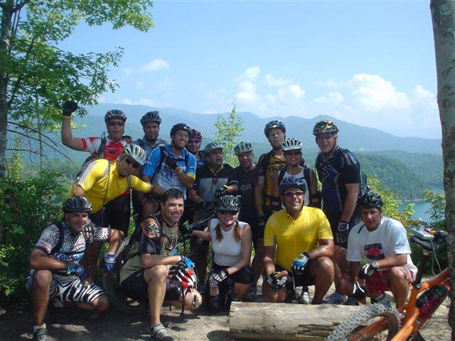 A group of enthusiastic mountain bikers poses for a photo at a scenic overlook. They are wearing colorful cycling gear and helmets, standing in front of a backdrop of mountains and a blue sky. Some of the riders are giving thumbs up or peace signs, showcasing their camaraderie and enjoyment of the sport. Tsali Mouse Branch Loop mountain bike trail.