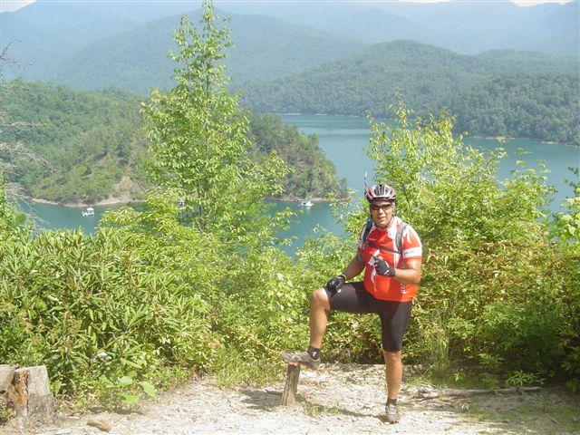 A cyclist in a red jersey and helmet stands on a hiking trail, posing with one leg raised on a small log. In the background, a scenic view of a lake surrounded by lush green hills and mountains is visible under a clear blue sky. Tsali Mouse Branch Loop mountain bike trail.