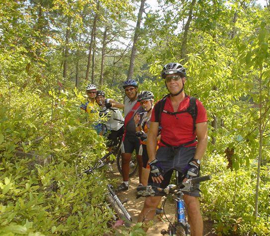 A group of five mountain bikers stopped on a trail surrounded by lush greenery and trees. They are wearing helmets and cycling gear, smiling and posing for the photo, with bicycles partially visible in the foreground. The scene captures a sunny day outdoors and conveys a sense of camaraderie and adventure. Tsali Right Loop mountain bike trail.