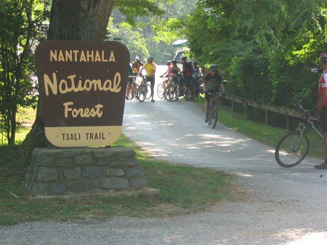 A sign for Nantahala National Forest marks the entrance to Tsali Trail, with a group of mountain bikers gathered nearby. The path is surrounded by lush greenery and trees, creating a scenic outdoor environment. Tsali Right Loop mountain bike trail.