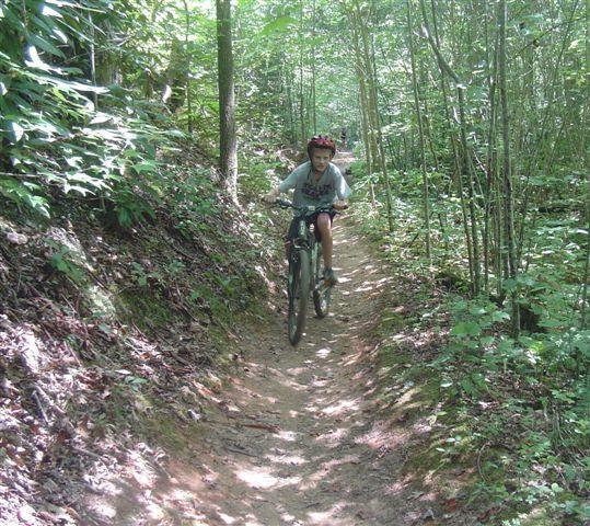 A young person riding a mountain bike along a narrow dirt trail in a lush green forest. The trail is surrounded by tall trees and dense foliage, creating a natural setting for outdoor biking. The cyclist is wearing a helmet and is focused on navigating the path. Tsali Thompson Loop mountain bike trail.