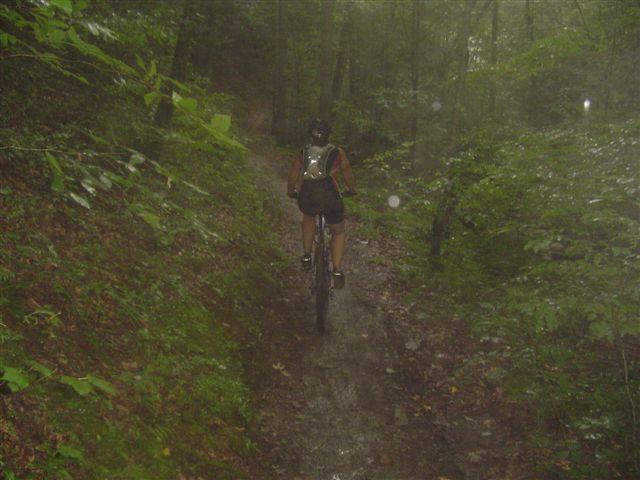A person riding a mountain bike on a narrow, muddy trail surrounded by dense greenery in a misty forest setting. Tsali Thompson Loop mountain bike trail.