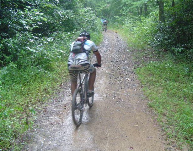 A cyclist riding on a muddy trail surrounded by lush greenery, with another cyclist visible in the background. Tsali Thompson Loop mountain bike trail.