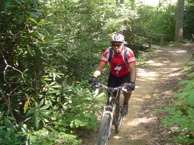 A mountain biker wearing a red jersey and helmet navigates a dirt path surrounded by lush green foliage. The cyclist is focused and pedaling uphill on a scenic trail in a wooded area. Tsali Thompson Loop mountain bike trail.