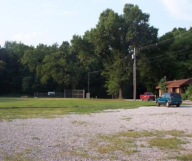 A grassy area with a gravel parking lot, featuring two parked vehicles—one green and one red. In the background, there is a baseball field surrounded by trees under a clear sky. A small building is visible to the right. Scales Lake Park Trails mountain bike trail.