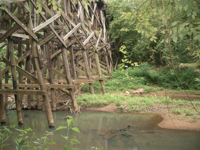 A dilapidated wooden structure stands over a shallow creek surrounded by lush greenery. The support beams are partially submerged in water, with patches of grass and earth visible along the banks. The surrounding trees and foliage create a natural, serene environment. Silver Comet Rail Trail mountain bike trail.